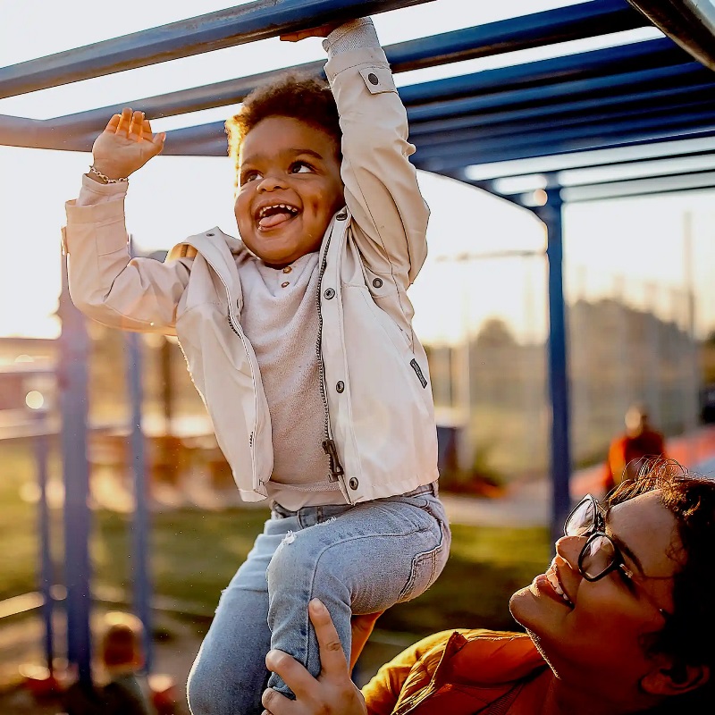 Photo of a mother smiling and holding her toddler up, while he plays on the monkey bars at a playground.